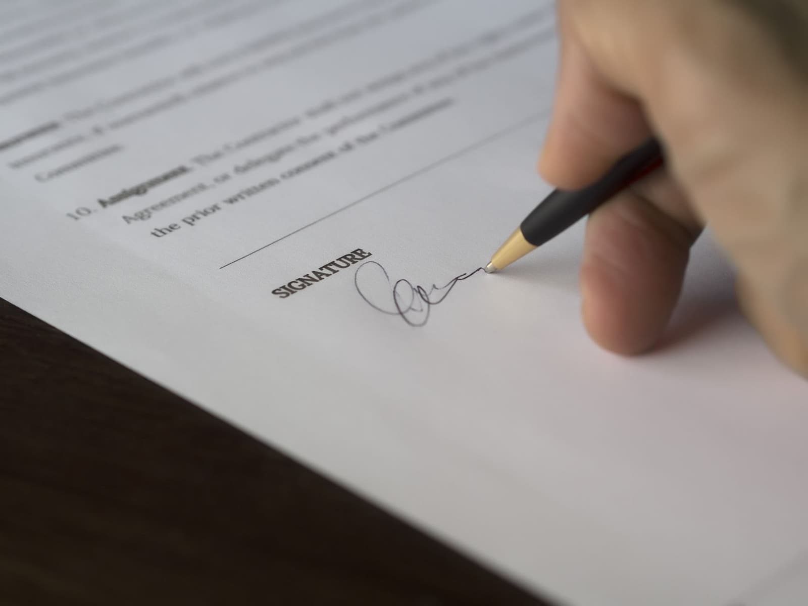 Una mano firma un contrato con bolígrafo sobre un escritorio de madera.