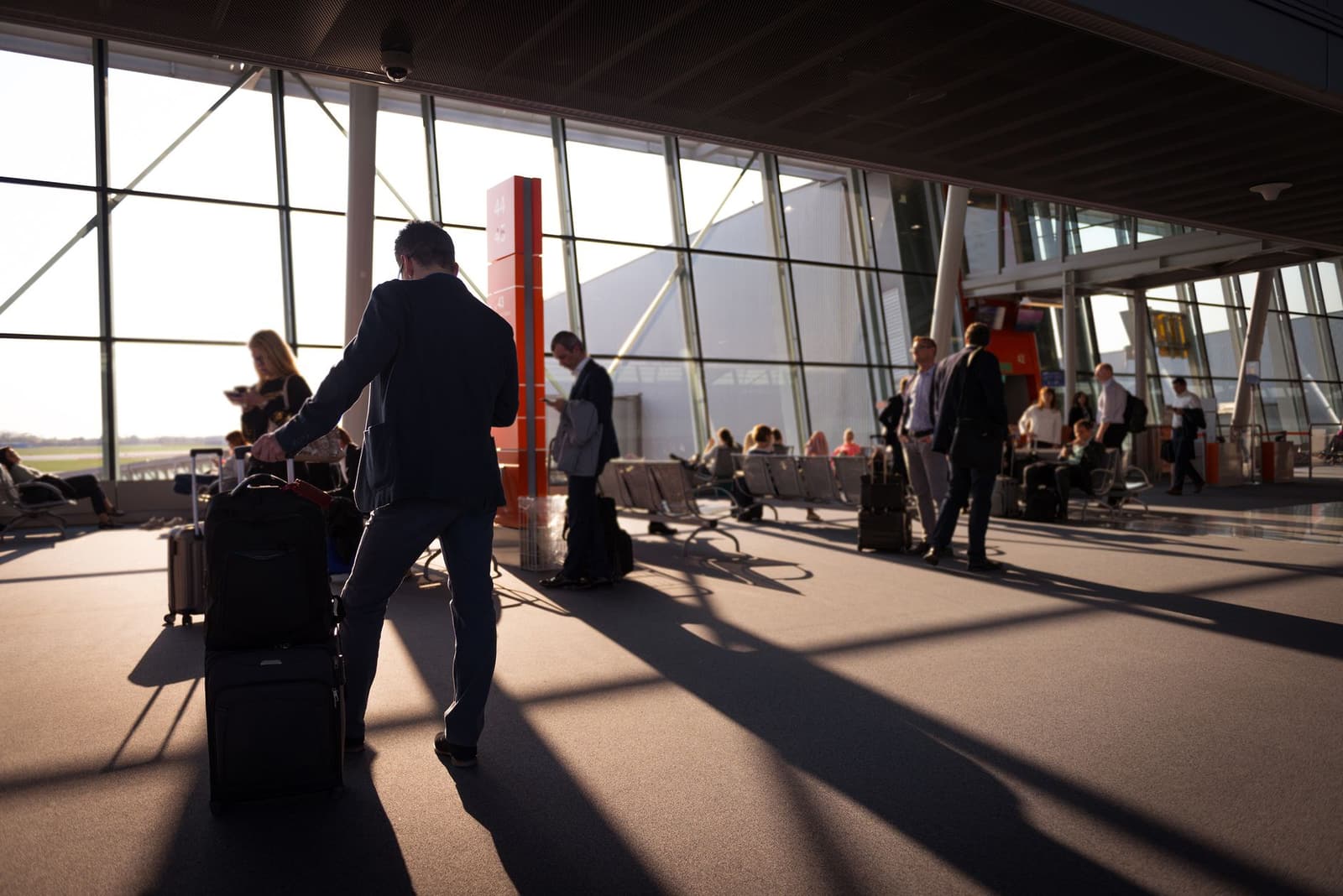 Pasajeros con equipaje cruzan a contraluz una sala de embarque de aeropuerto.